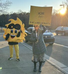 Sun mascot and teacher with sign: "Honk if you carpooled today"